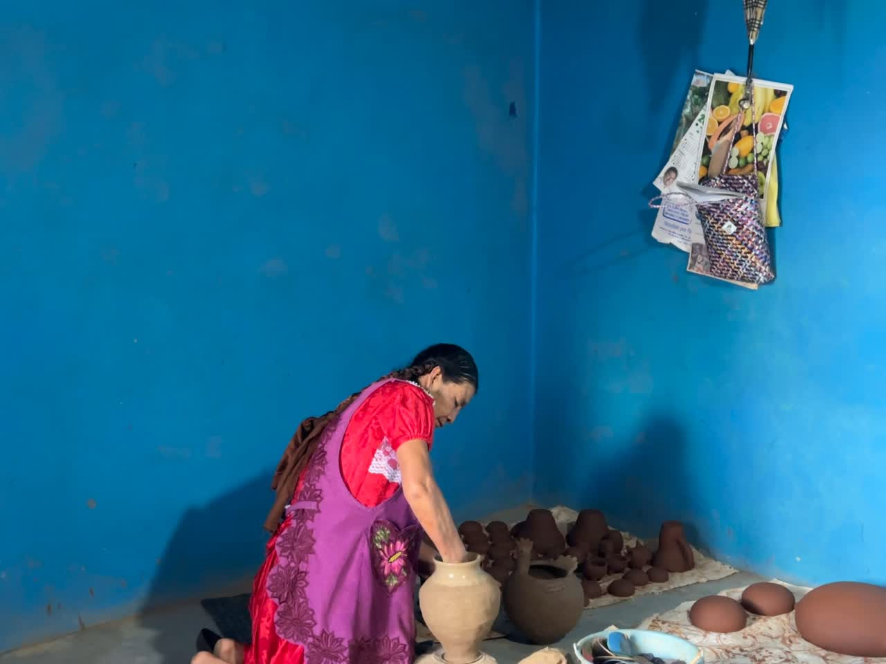 A potter shaping a vessel by hand against a vivid blue wall in Oaxaca